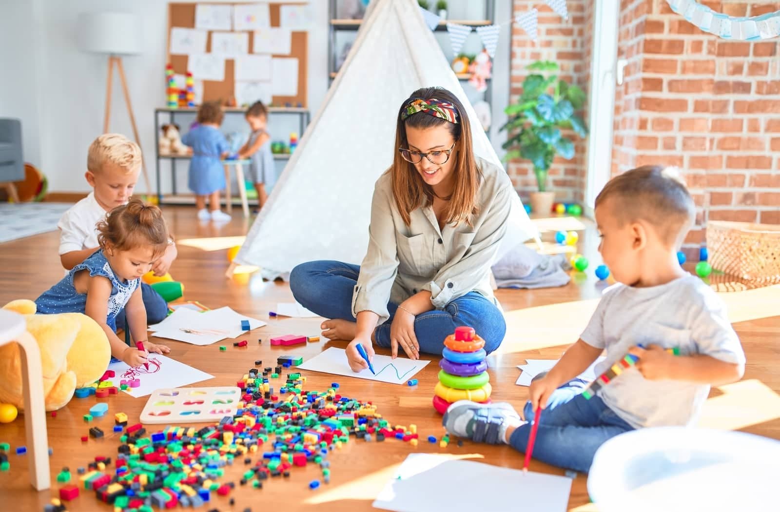 A caregiver sitting on the floor drawing with toddlers in a bright classroom
